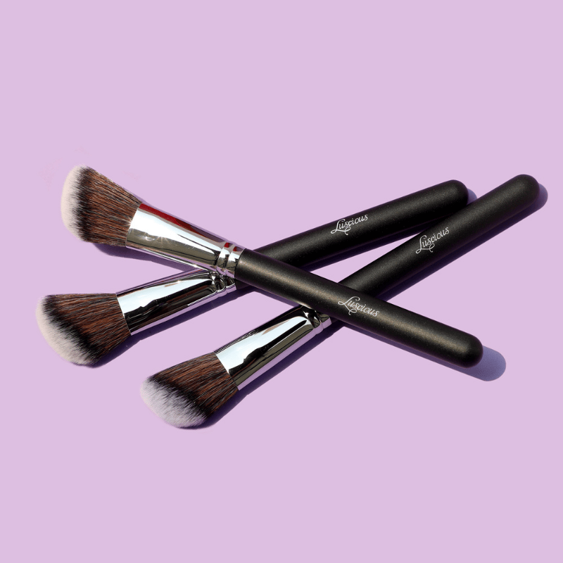 Luscious Cosmetics makeup brushes with black handles and wooden bristles on a light purple background.