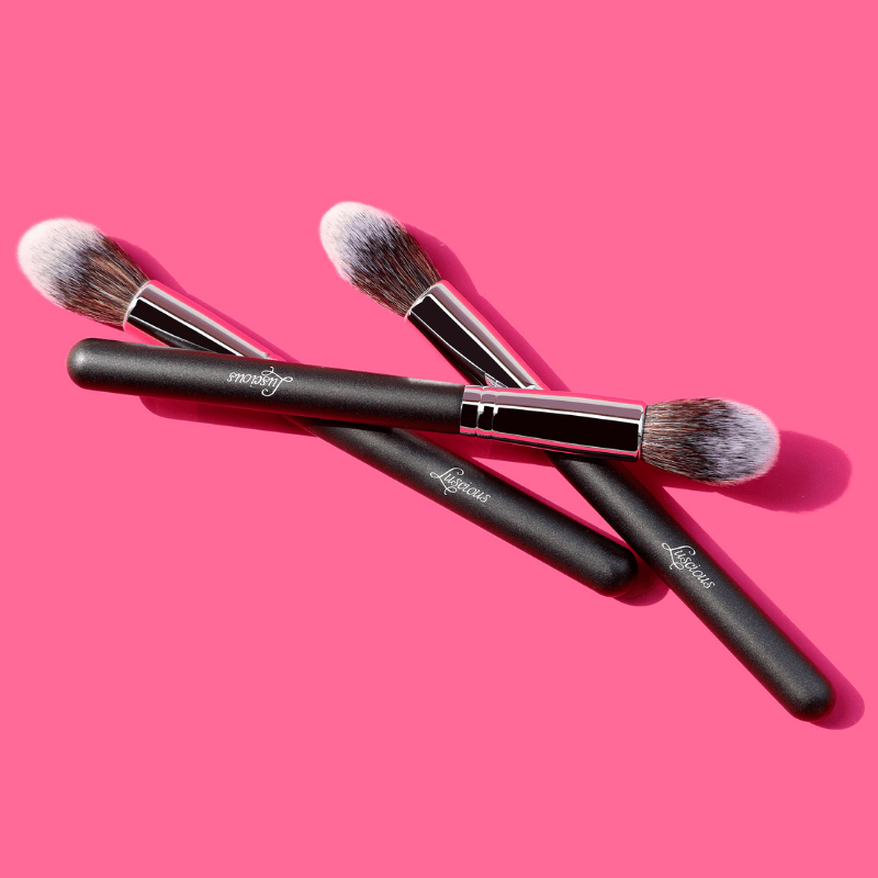 Luscious Cosmetics blush highlight brush with black handle and silver accents on a pink background.