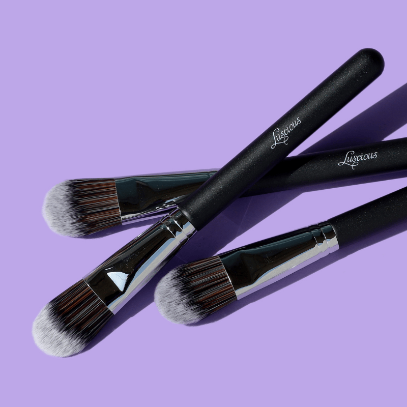 Luscious Cosmetics foundation makeup brush with black handle and silver ferrule on a purple background.
