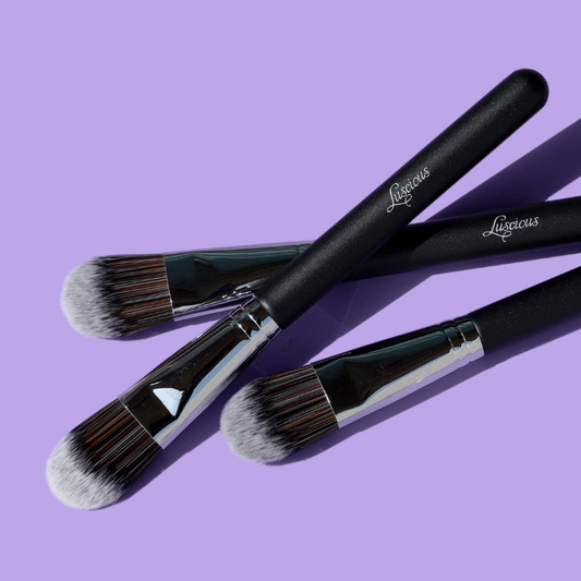 Luscious Cosmetics foundation makeup brush with black handle and silver ferrule on a purple background.