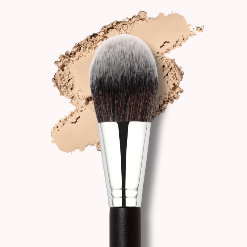 Luscious Cosmetics all-over powder brush with a flat, round synthetic head and silver metal handle, shown on a beige powder swatch.