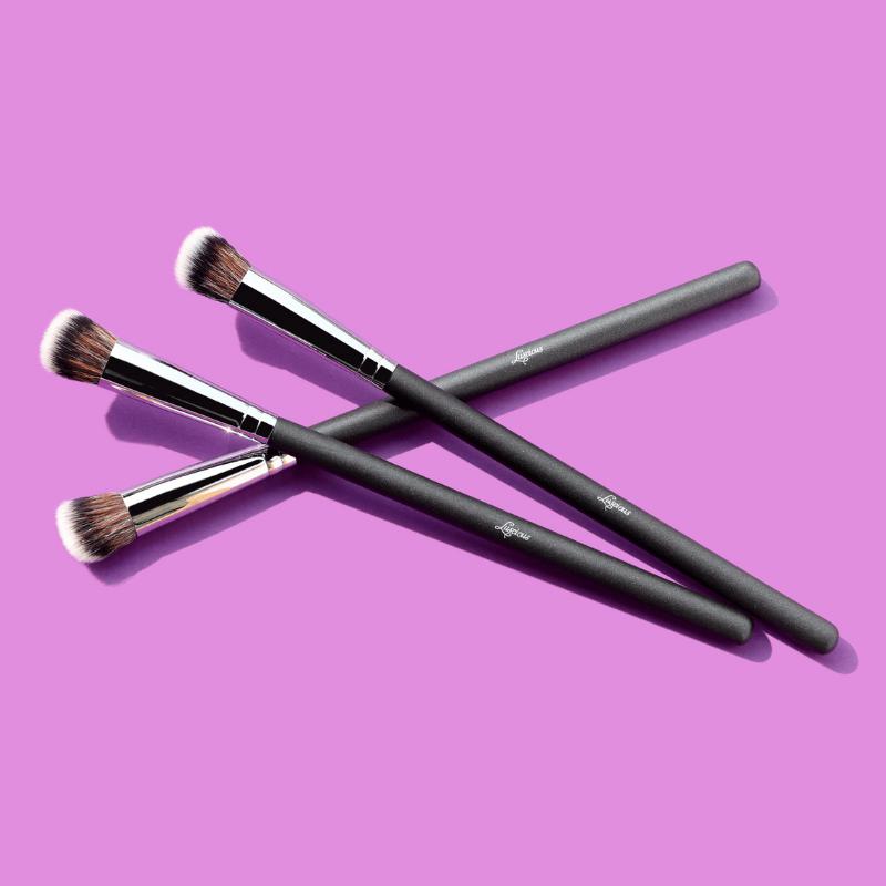 Luscious Cosmetics makeup brushes with black handles and brown bristles on a purple background.