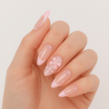A hand wearing glossy pink press-on nails by Luscious Cosmetics, decorated with a French-style design and pearl bow detail.
