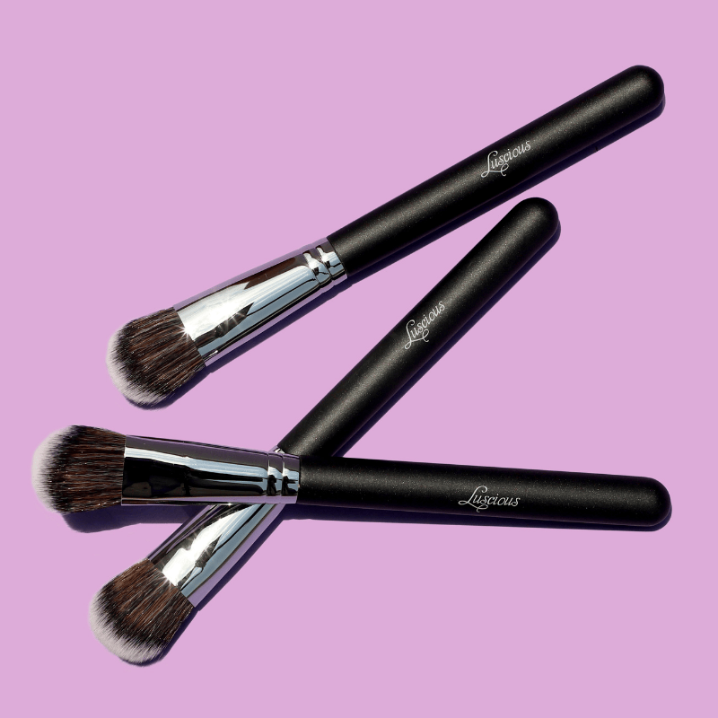 Luscious Cosmetics makeup brush set with black handles and silver ferrules on a pink background.